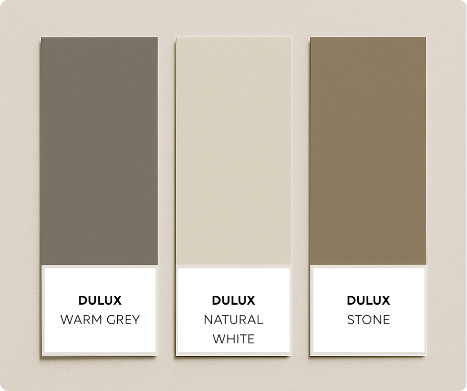 Three Dulux paint swatches are displayed vertically on a pale textured background. The colours shown are Warm Grey (medium grey), Natural White (creamy off-white), and Taupe Stone (taupe brown-grey), each labeled beneath the swatch.